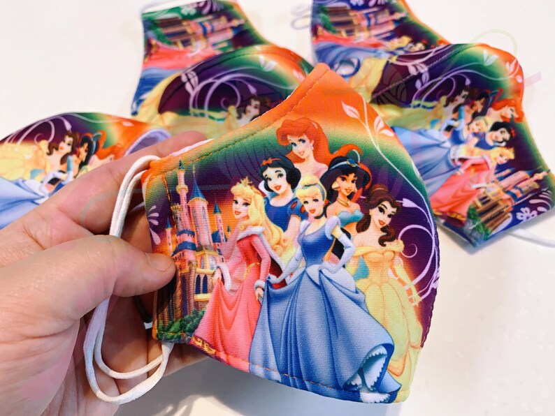 May include: A colourful face mask featuring a group of Disney princesses, including Cinderella, Snow White, Belle, and Ariel. The mask has a purple, orange, and green background with a castle in the background.