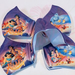 May include: Four purple fabric face masks with different Disney Aladdin characters printed on them. The masks feature Aladdin, Jasmine, Abu, and Genie. The masks are folded and stacked on top of each other.