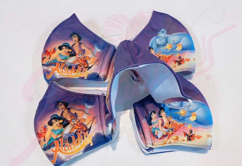 May include: Four purple fabric face masks with different Disney Aladdin characters printed on them. The masks feature Aladdin, Jasmine, Abu, and Genie. The masks are folded and stacked on top of each other.