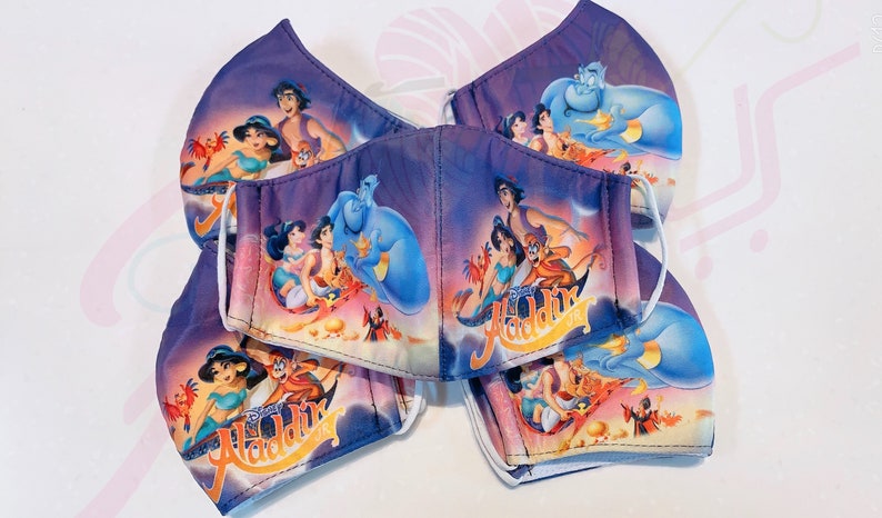 May include: A set of five purple and blue face masks featuring a colourful image of the Disney characters from Aladdin, including Aladdin, Jasmine, Abu, and Genie. The masks have white elastic ear loops.
