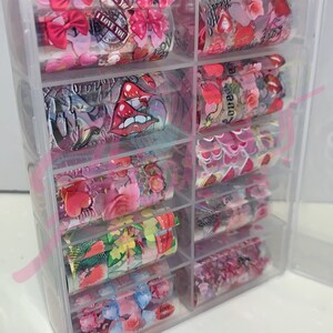 以下が含まれることがあります： ハート、リボン、唇、花、そして「I Love You」や「Love Love」などの言葉がデザインされたネイルアート転写ホイルのロールが、透明なプラスチック製の収納ボックスに入っています。
