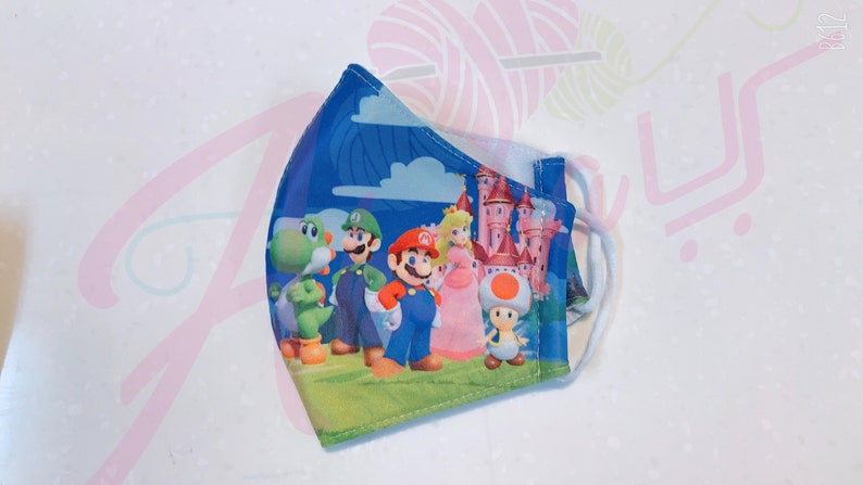 May include: A blue face mask with a colorful print of Mario, Luigi, Peach, Yoshi, and Toad. The characters are standing in front of a pink castle.