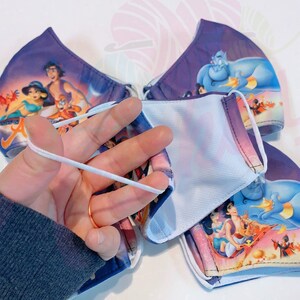 May include: Four reusable face masks with a purple background and a colourful image of characters from the Disney film Aladdin. The masks have white elastic ear loops.