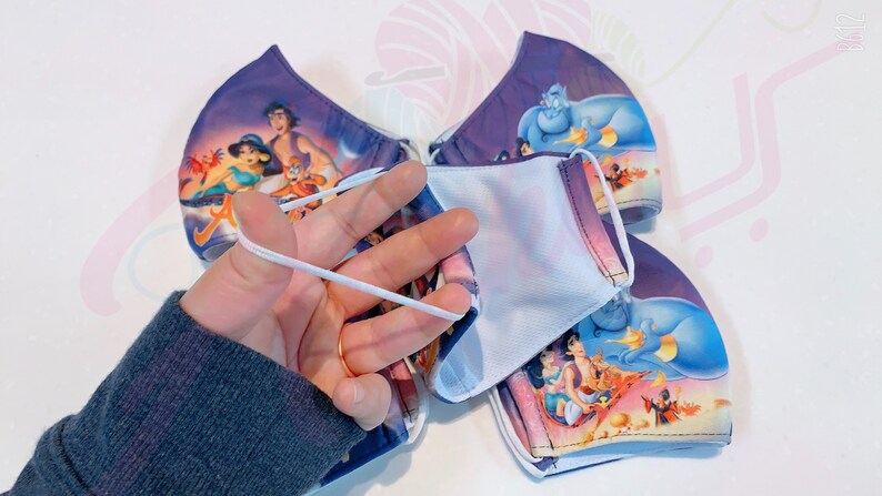 May include: Four reusable face masks with a purple background and a colourful image of characters from the Disney film Aladdin. The masks have white elastic ear loops.