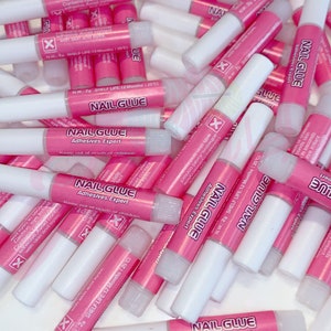Op de afbeelding: Een stapel roze en witte nagellijm tubes. De tubes zijn gelabeld met "Nail Glue Adhesives Expert" en hebben een waarschuwingssticker met de tekst "Let op: irriterend voor de ogen Bevat cyanoacrylaatlijm Vermijd contact met huid en ogen."