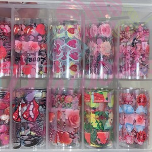 以下が含まれることがあります： 透明なプラスチック容器に入った10枚のネイルアート転写ホイルのセット。ホイルには、ハート、花、唇、そして「love」という言葉など、さまざまなデザインが施されています。