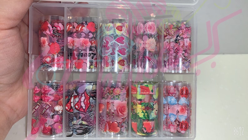 以下が含まれることがあります： 透明なプラスチック容器に入った10枚のネイルアート転写ホイルのセット。ホイルには、ハート、花、唇、そして「love」という言葉など、さまざまなデザインが施されています。