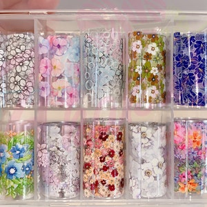 May include: A clear plastic storage box with 12 rolls of floral patterned nail art transfer foil. The foil is in various colors and patterns, including pink, blue, green, and white.