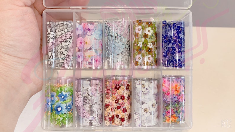 May include: A clear plastic storage box with 12 rolls of floral patterned nail art transfer foil. The foil is in various colors and patterns, including pink, blue, green, and white.