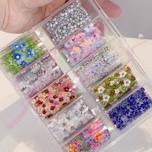 May include: A clear plastic box containing ten rolls of nail art foil. Each roll features a different floral design with various colors, including blue, pink, green, and white.