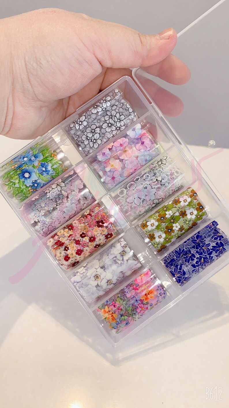 May include: A clear plastic box containing ten rolls of nail art foil. Each roll features a different floral design with various colors, including blue, pink, green, and white.