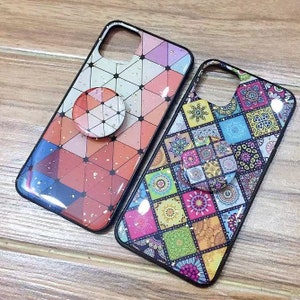 May include: Two phone cases with a geometric design. One case is red, white, and black with gold glitter. The other case is multi-colored with a patchwork design. Both cases have a pop socket.