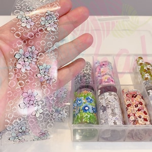 May include: A clear plastic box filled with rolls of nail art transfer foil. The foil has various floral patterns in different colors, including pink, blue, green, and white. One roll of foil is being held in a hand, showing the floral pattern more clearly.