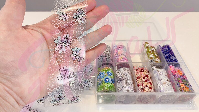 May include: A clear plastic box filled with rolls of nail art transfer foil. The foil has various floral patterns in different colors, including pink, blue, green, and white. One roll of foil is being held in a hand, showing the floral pattern more clearly.