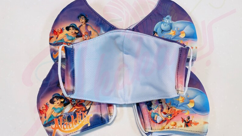 May include: A set of four purple and blue face masks featuring a printed image of Aladdin and Genie from the Disney film Aladdin. The masks have white elastic ear loops and a white inner layer.