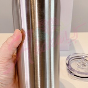 Puede incluir: Un vaso de acero inoxidable con acabado cepillado. El vaso se sostiene en una mano y parece ser alto y cil&iacute;ndrico.
