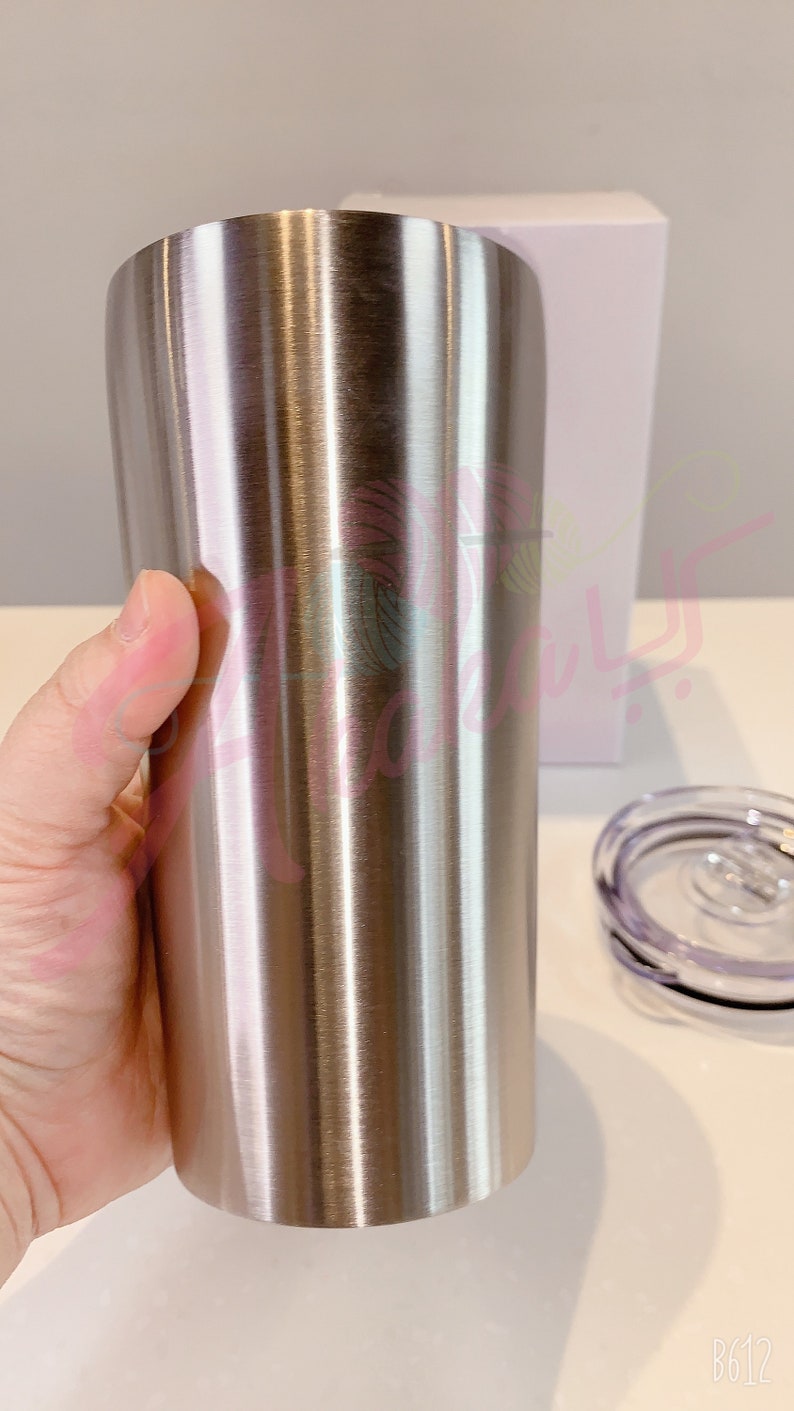 Puede incluir: Un vaso de acero inoxidable con acabado cepillado. El vaso se sostiene en una mano y parece ser alto y cil&iacute;ndrico.
