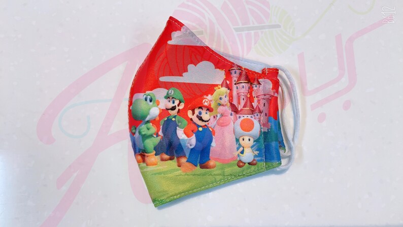 May include: A red, white, and blue face mask with a print of Mario, Luigi, Peach, Yoshi, and Toad. The mask features a castle and clouds in the background.