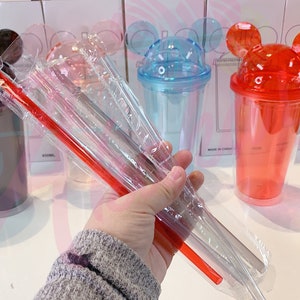 May include: Set of six clear plastic tumblers with Mickey Mouse ears on the lid. The tumblers are in various colors, including green, black, blue, red, and clear. The set also includes three clear plastic straws and one red plastic straw.