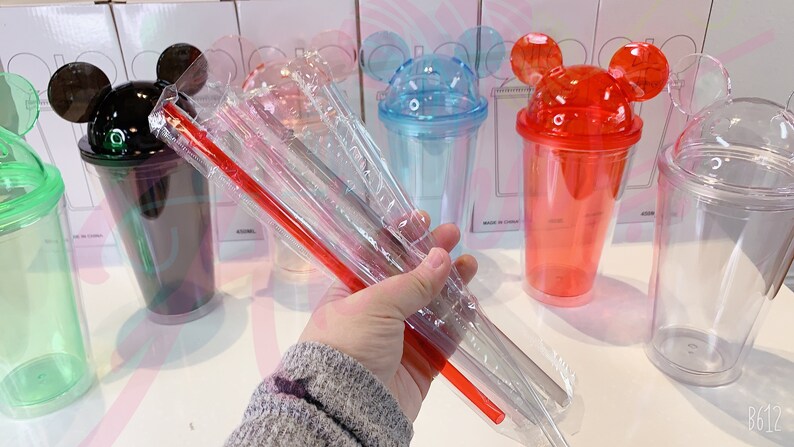 May include: Set of six clear plastic tumblers with Mickey Mouse ears on the lid. The tumblers are in various colors, including green, black, blue, red, and clear. The set also includes three clear plastic straws and one red plastic straw.