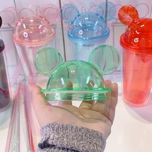 May include: A set of six clear plastic cups with Mickey Mouse ears on the lids. The cups are in various colors, including black, pink, blue, red, green, and clear. The cups are all the same size and shape.