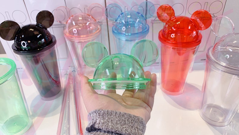 May include: A set of six clear plastic cups with Mickey Mouse ears on the lids. The cups are in various colors, including black, pink, blue, red, green, and clear. The cups are all the same size and shape.