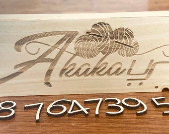 May include: A wooden sign with the word "Ahakal" engraved in cursive, with two yarn balls above the word. Below the sign are the numbers 08 764 739 58.