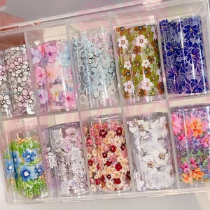 May include: A clear plastic storage box with 10 rolls of floral patterned nail art transfer foil. The foil is in various colors and patterns, including white, pink, blue, and green.