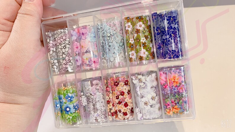 May include: A clear plastic storage box with 10 rolls of floral patterned nail art transfer foil. The foil is in various colors and patterns, including white, pink, blue, and green.
