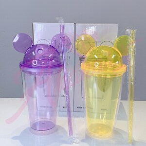 May include: Two clear plastic cups with lids and straws. The cups have a Mickey Mouse ear design on the lid. One cup is purple and the other is yellow. The cups are labeled "450ML" and "MADE IN CHINA".