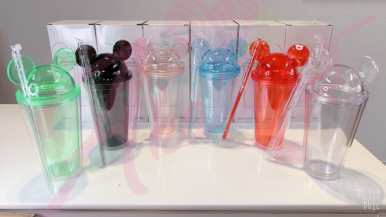 May include: Six clear plastic cups with Mickey Mouse ears and lids. The cups are in different colors: green, black, pink, blue, red, and clear. Each cup comes with a straw.