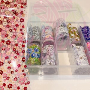 May include: A clear plastic storage box with nine rolls of nail art transfer foil. The foil has various floral patterns in different colors, including pink, blue, green, and purple.