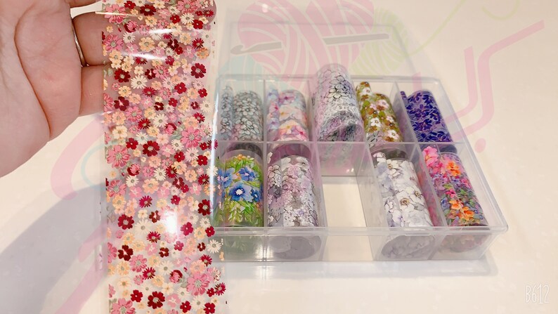 May include: A clear plastic storage box with nine rolls of nail art transfer foil. The foil has various floral patterns in different colors, including pink, blue, green, and purple.