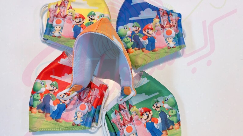 May include: Four colorful face masks featuring characters from the Super Mario Bros. video game franchise. The masks are made of fabric and have elastic ear loops. The characters on the masks include Mario, Luigi, Peach, Toad, and Yoshi.
