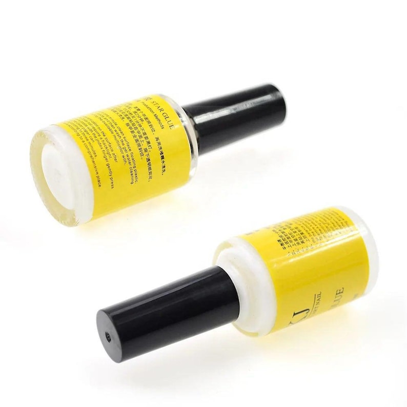 May include: Two bottles of nail glue are displayed against a white background. The bottles have a clear base with a yellow label and black cap. The label includes text in multiple languages. The bottles are angled, showing the product and label details.