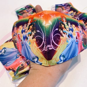May include: A colourful face mask featuring a design of Disney princesses, including Cinderella, Belle, and Snow White. The mask is made of fabric and has a purple, orange, and green background.