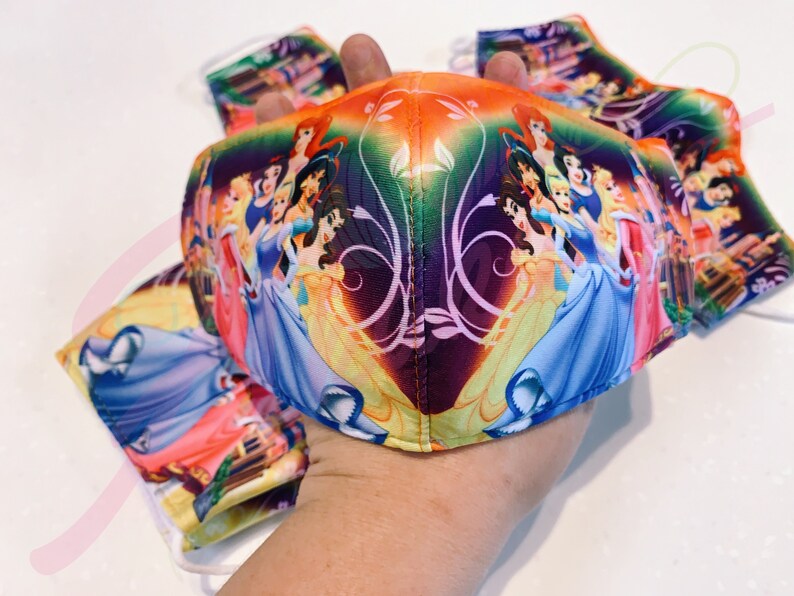 May include: A colourful face mask featuring a design of Disney princesses, including Cinderella, Belle, and Snow White. The mask is made of fabric and has a purple, orange, and green background.