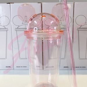 May include: Pink plastic tumbler with a lid and a straw. The tumbler has a Mickey Mouse ear design and a measurement scale on the side. The tumbler is 450 ml.