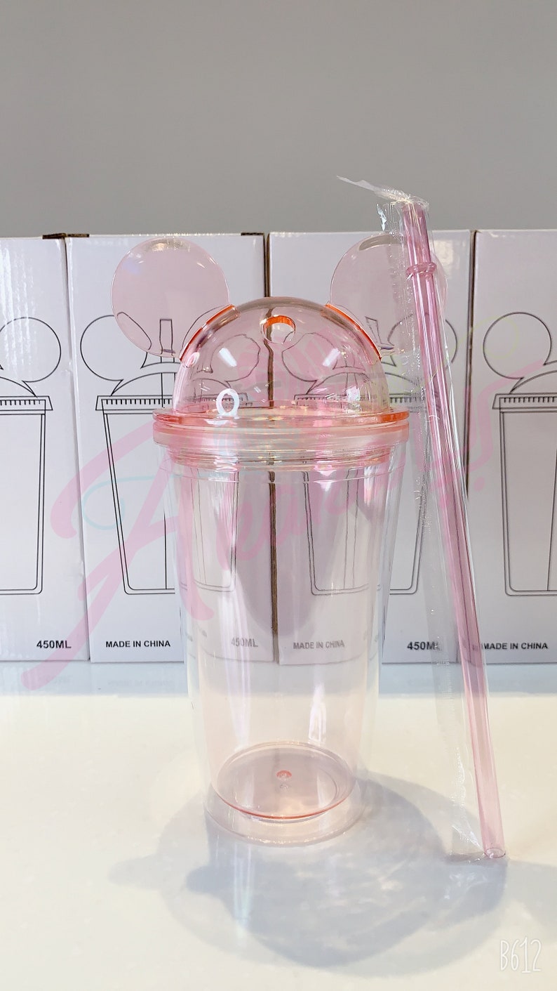 May include: Pink plastic tumbler with a lid and a straw. The tumbler has a Mickey Mouse ear design and a measurement scale on the side. The tumbler is 450 ml.