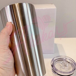 Puede incluir: Un vaso de acero inoxidable con acabado cepillado. El vaso se sostiene en una mano y est&aacute; al lado de una caja blanca y una tapa transparente.