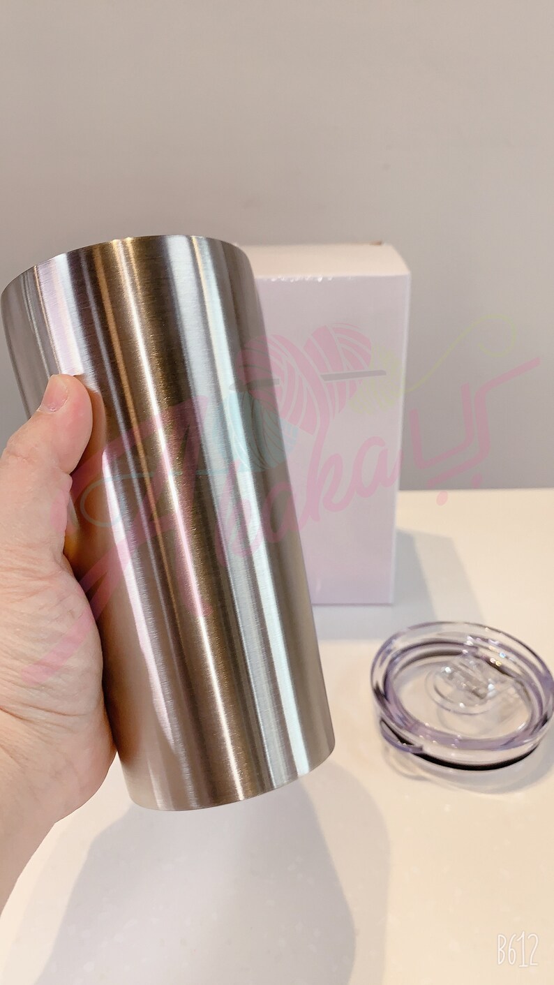 Puede incluir: Un vaso de acero inoxidable con acabado cepillado. El vaso se sostiene en una mano y est&aacute; al lado de una caja blanca y una tapa transparente.