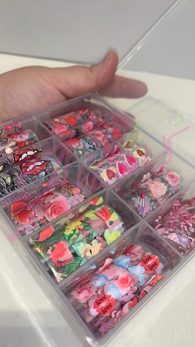 以下が含まれることがあります： 透明なプラスチック製の箱に、ネイルアート用のホイルのロールが詰められています。ホイルには、ハート、花、唇、そして「love」という言葉など、さまざまなデザインが施されています。