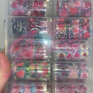 以下が含まれることがあります： 透明なプラスチック容器に、ネイルアート転写ホイルが10ロール入っています。ホイルには、ハート、花、そして「love」や「sweet love」の文字が書かれた、様々なピンクと白のデザインが施されています。