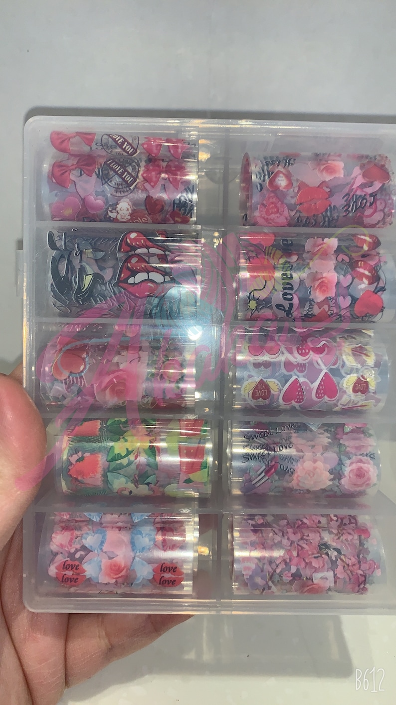 以下が含まれることがあります： 透明なプラスチック容器に、ネイルアート転写ホイルが10ロール入っています。ホイルには、ハート、花、そして「love」や「sweet love」の文字が書かれた、様々なピンクと白のデザインが施されています。