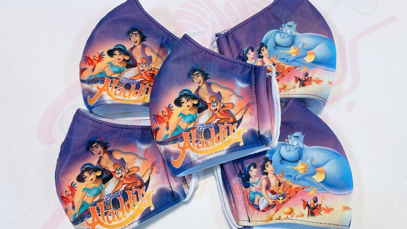 May include: Five purple face masks featuring a printed image of Aladdin and Jasmine on a magic carpet. The masks have white elastic ear loops.