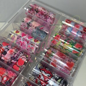 以下が含まれることがあります： カラフルなネイルアートホイルのロールが詰まった透明なプラスチック製の収納ボックス。ホイルには、ハート、花、抽象的なパターンなど、さまざまなデザインが施されています。