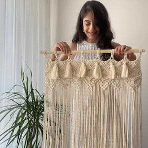 Könnte beinhalten: Cremefarbener Makramee-Wandbehang mit einem Holzstab. Das Makramee hat ein geometrisches Design mit Quasten. Das Stück wird von einer Person in einem weißen Oberteil gehalten. Im Hintergrund ist eine grüne Pflanze zu sehen.