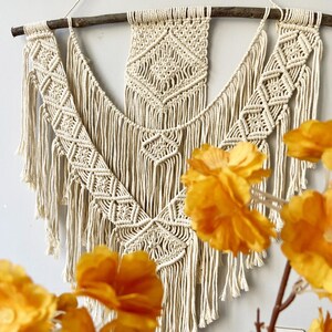 Puede incluir: Tapiz de pared de macramé color crema con un patrón de diamantes geométricos y flecos. El macramé está suspendido de una rama de madera natural. Flores amarillas en primer plano.