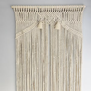 Könnte beinhalten: Ein cremefarbener Makramee-Wandbehang mit einem Holzstab an der Oberseite, komplizierten Knotenmustern und zwei dekorativen Quasten. Das Design umfasst lange, vertikal hängende Schnüre mit kleinen Knoten, die über die gesamte Länge verteilt sind.