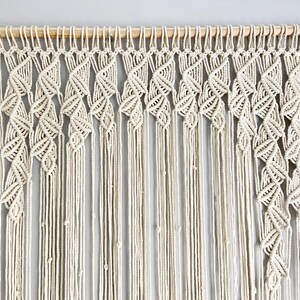May include: Cream-colored macrame wall hanging. The design features a wooden dowel at the top, with intricate knotted patterns forming a cascading, leaf-like design. Long, vertical strands hang below, creating a textured, bohemian aesthetic.
